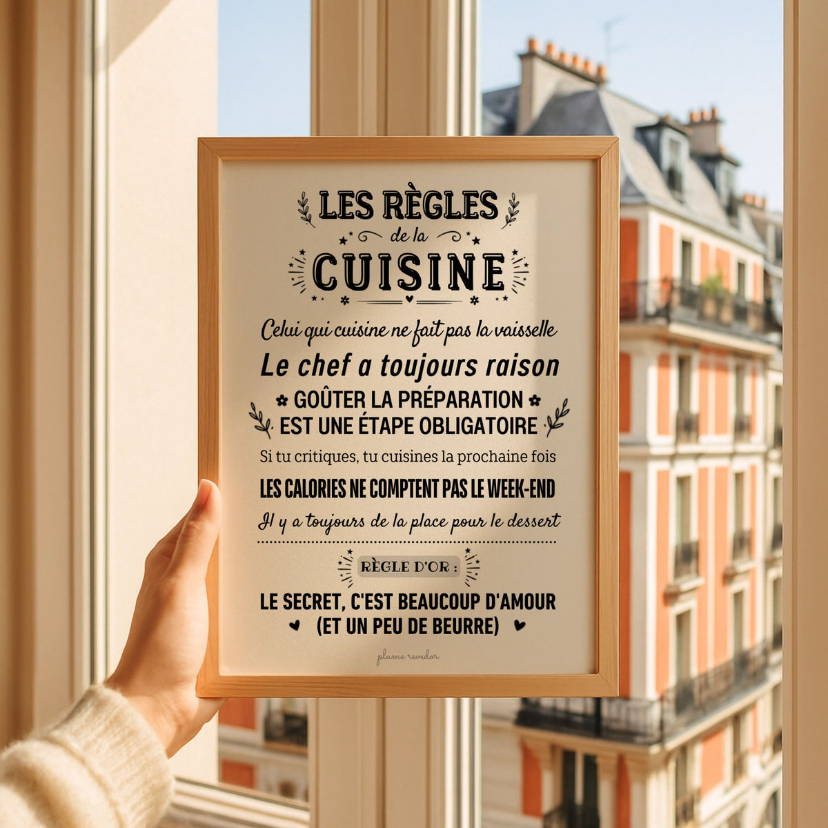 Affiche « Les règles de la cuisine » dans un cadre en bois, tenue devant une fenêtre donnant sur des immeubles.