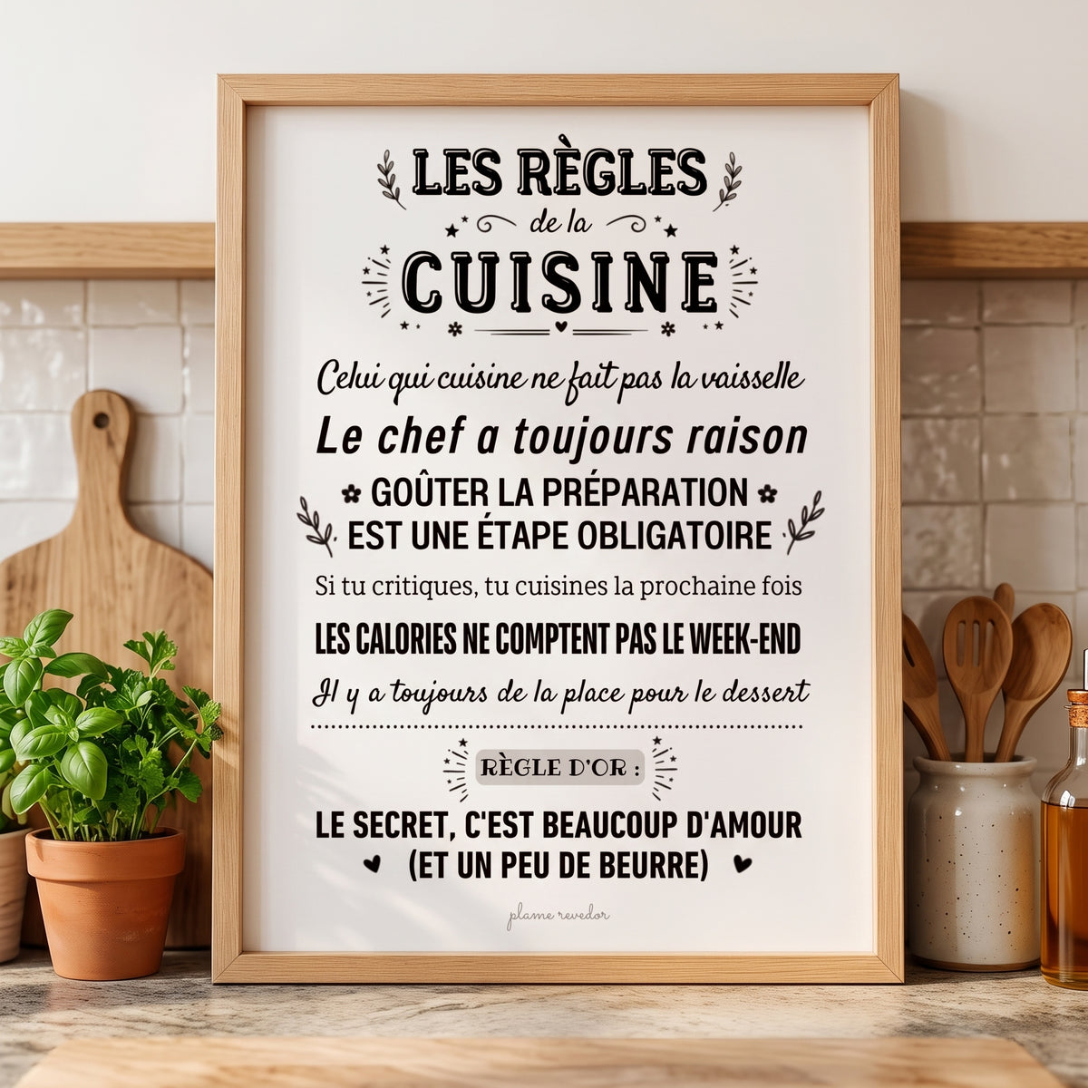 Affiche « Les règles de la cuisine » dans un cadre en bois, sur un plan de travail avec des herbes et des ustensiles.