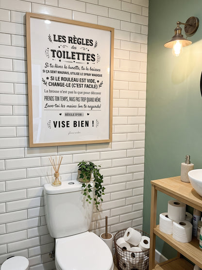 Affiche « Les règles des toilettes » dans un cadre clair, au-dessus des WC sur un mur en carrelage blanc.