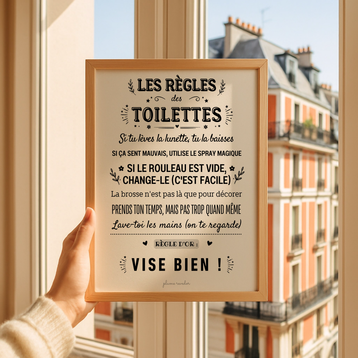 Affiche « Les règles des toilettes » dans un cadre en bois, tenue devant une fenêtre donnant sur la ville.