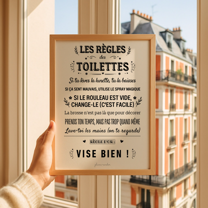 Affiche « Les règles des toilettes » dans un cadre en bois, tenue devant une fenêtre donnant sur la ville.