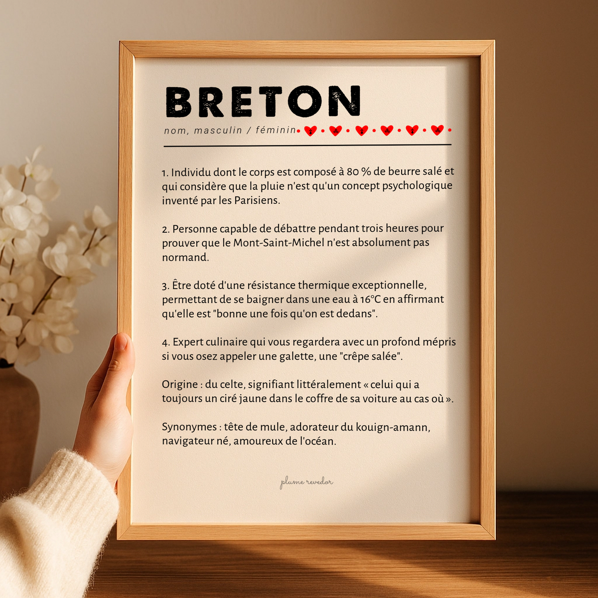 Affiche humoristique définition Breton dans un cadre en bois, tenue au-dessus d’un meuble près de fleurs blanches.